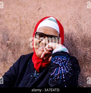 MARRAKECH, MAROC - 16 MAI 2024 femme traditionnelle en bleu avec foulard rouge avec dos contre un mur dans le marché du souk dans la médina Banque D'Images