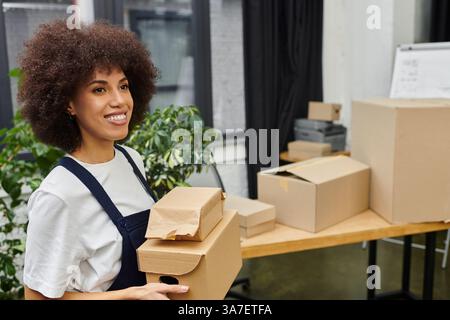 Une jeune femme sourit alors qu'elle porte des boîtes emballées tout en se préparant à s'installer dans son nouvel espace. Banque D'Images