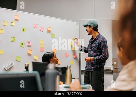 Souriant jeune homme collant une note adhésive sur le tableau blanc tout en discutant du projet en classe avec des amis à l'université Banque D'Images