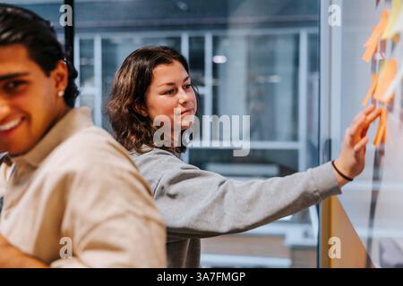 Jeune femme collant une note adhésive sur le tableau blanc dans la salle de classe au collège Banque D'Images
