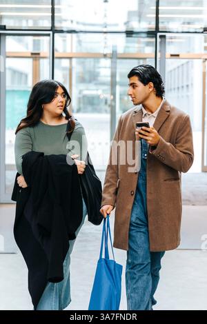 Vue avant du jeune homme tenant sac et téléphone intelligent parlant avec une amie en marchant dans le campus universitaire Banque D'Images