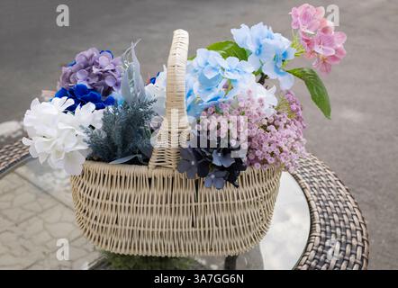 bouquet luxuriant de fleurs artificielles d'hortensia multicolores dans un beau panier en osier sur une table en verre. Banque D'Images