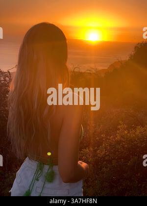 Une jeune femme aux cheveux longs se tient sur une colline, regardant le coucher du soleil. Banque D'Images