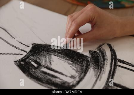 Femme dessinant image avec crayon de cire noir sur papier à table dans l'atelier de l'artiste, gros plan Banque D'Images