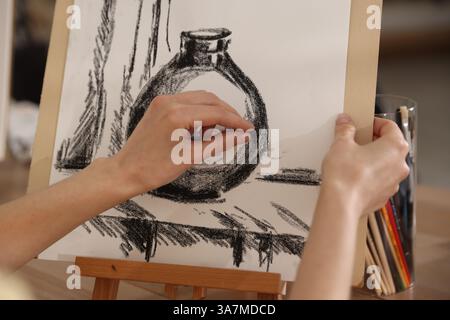 Femme dessinant image avec crayon de cire noir sur chevalet à la table en bois dans l'atelier de l'artiste, gros plan Banque D'Images