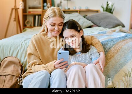 Femme et enfant assis sur le sol, souriant au smartphone. Chambre confortable avec couvertures et oreillers moelleux, les deux portent des vêtements décontractés Banque D'Images