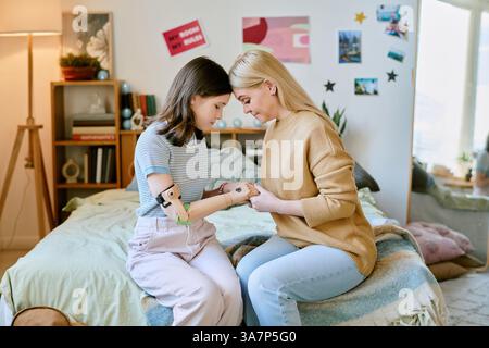 Deux femmes caucasiennes assises sur le lit, se tenant les mains dans une chambre confortable et bien décorée, fille portant un bras prothétique et souriant tout en s'engageant avec la mère Banque D'Images