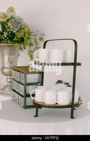 Mugs blancs disposés sur un stand à deux niveaux à côté de fleurs décoratives dans un espace intérieur serein. Banque D'Images