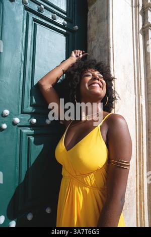 Belle femme noire avec des cheveux de corbeau portant une robe jaune posant pour la photo contre la grande porte verte de l'église. Heureux et souriant Banque D'Images
