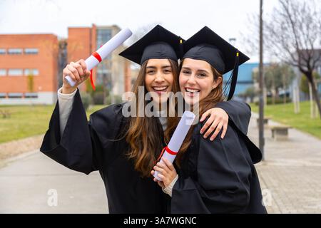 Deux étudiantes portant des robes et des casquettes de remise des diplômes sont heureuses de tenir leurs diplômes et de s'embrasser le jour de la remise des diplômes Banque D'Images