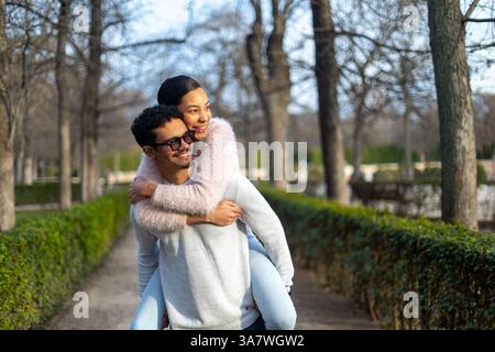 Jeune homme donnant piggyback ride à sa petite amie tout en marchant dans un parc, profitant d'un moment romantique ensemble Banque D'Images