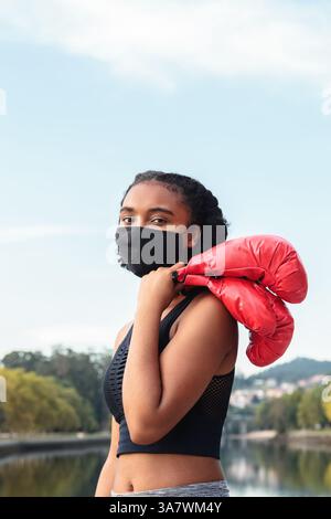Boxeur féminin afro-américain sérieux dans le masque de protection et gants rouges pendant l'entraînement sur la rive du lac dans le parc Banque D'Images