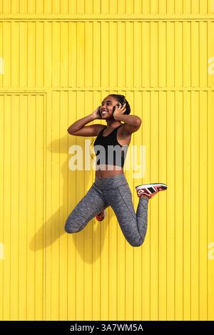 Pleine longueur de jeune sportif afro-américain joyeux en activewear sautez contre le mur jaune et écoutez de la musique dans un casque pendant le train Banque D'Images