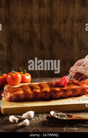 Crop anonyme chef étalant la sauce barbecue sur les côtes de porc crues tout en cuisinant à table avec divers ingrédients Banque D'Images
