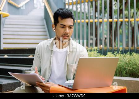 Jeune indépendant asiatique portant des écouteurs et tenant des documents fonctionne à distance sur un ordinateur portable tout en se relaxant sur un banc extérieur dans un cadre urbain Banque D'Images