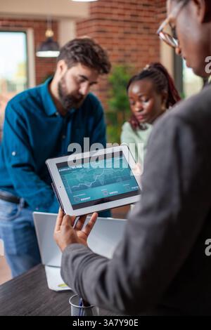 Black Analyst utilise la tablette pour évaluer les rapports d'entreprise tandis que ses collègues travaillent ensemble dans le bureau de démarrage. Le responsable afro-américain examine les progrès de l'entreprise sur les appareils numériques, les collègues discutent à proximité. Banque D'Images