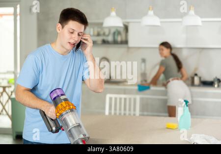 Jeune homme positif parlant au téléphone portable et aspirant le sol de la cuisine Banque D'Images