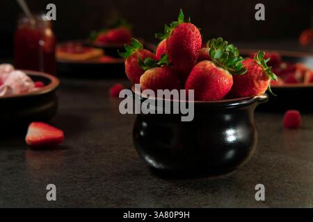 Vue de face d'un pot en céramique noir rempli de fraises au premier plan sur un fond d'assiettes avec des desserts aux fraises Banque D'Images