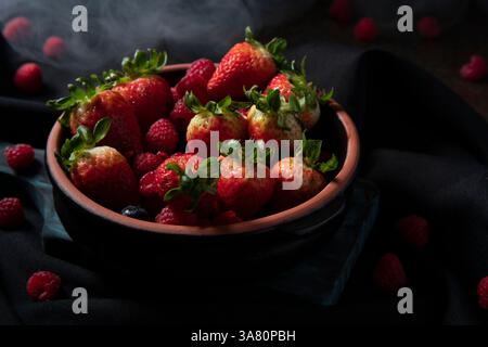 Gros plan d'un plateau en céramique noir rempli de baies rouges, de fraises, de framboises et de bleuets sur une planche de bois foncé sur une table noire avec une râpe Banque D'Images