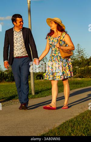 un couple souriant profite d'une promenade décontractée à l'extérieur, se tenant la main sous le ciel ensoleillé lumineux, mettant en valeur l'amour, le bonheur et la mode estivale dynamique dans un Banque D'Images