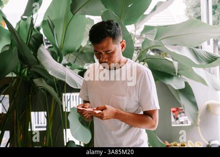 Un homme asiatique contemplatif dans un t-shirt blanc se tient au milieu de plantes tropicales luxuriantes, examinant attentivement un petit morceau de papier. Il semble concentré et th Banque D'Images