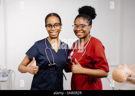 Deux femmes afro-américaines heureuses médecins portant des gommages colorés et des stéthoscopes faisant des gestes avec les pouces vers le haut dans une chambre d'hôpital Banque D'Images