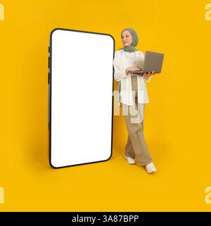 Souriant jolie fille musulmane caucasienne en hijab avec ordinateur portable regardant un énorme smartphone avec un écran blanc vide maquette. Fond jaune. Banque D'Images