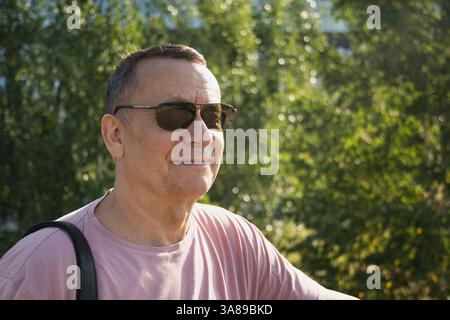 Portrait d'un homme adulte souriant portant des lunettes de soleil et un t-shirt rose au parc d'été. À l'extérieur. Gros plan. Banque D'Images