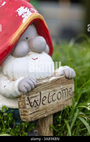 Gnome de jardin fantaisiste avec un chapeau rouge et un panneau en bois 'Bienvenue' entouré d'herbe verte luxuriante, créant une atmosphère charmante et accueillante. Banque D'Images