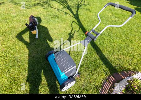 Ombre d'une personne debout près d'une tondeuse à gazon sur une pelouse verte pendant une journée ensoleillée, suggérant des soins de jardin et une activité de plein air. Banque D'Images