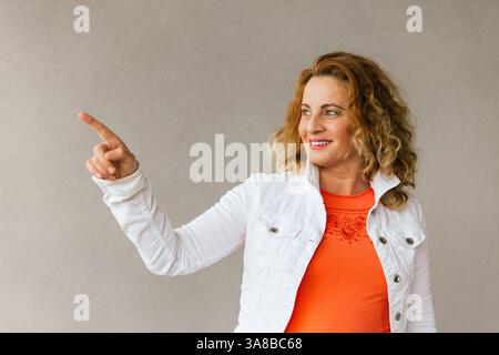 Femme d'âge moyen dans un haut orange et veste blanche pointant sur le côté avec un sourire joyeux, debout sur un fond neutre dans un a confiant Banque D'Images
