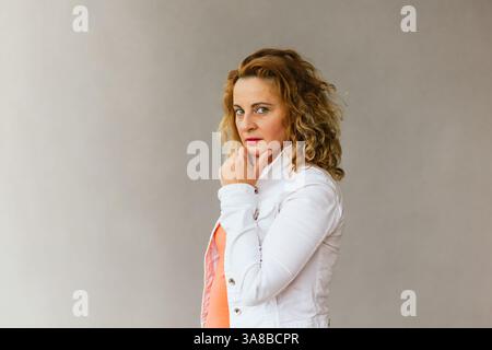 Femme d'âge moyen en haut orange et veste blanche debout dans le profil avec la main sur le menton, transmettant une humeur sérieuse et réfléchissante contre un dos neutre Banque D'Images