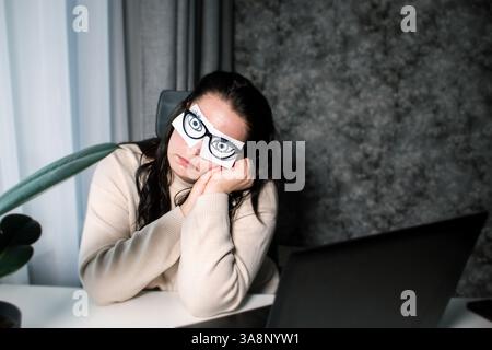 Femme d'affaires fatiguée avec des yeux peints sur des notes adhésives ayant la fatigue et des maux de tête pendant le travail au bureau. Sentiment féminin stressé et épuisé de Banque D'Images