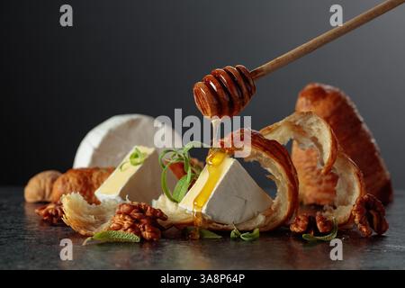 Camembert crémeux avec miel doré, croissants feuilletés et noix croquantes sur une table en pierre noire. Un mélange parfait d'élégance et de plaisir. Banque D'Images