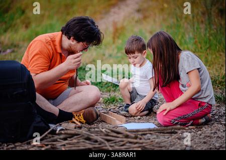 Un père et ses enfants se livrent à une activité de plein air pratique, apprenant ensemble des techniques de survie. Le cadre suggère un camping, un scoutisme ou un bushc Banque D'Images