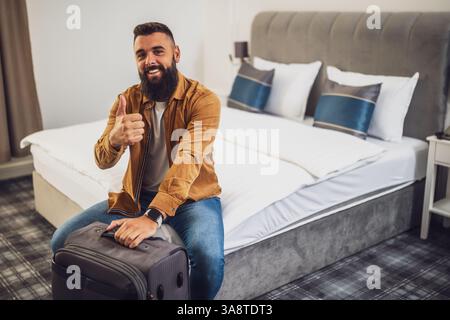 Un homme barbu joyeux est assis sur le lit dans la chambre d'hôtel et montre les pouces vers le haut. Il tient sa valise. Banque D'Images