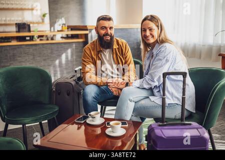 Heureux couple marié est arrivé à l'hôtel. Ils sont en vacances, assis dans le hall avec des valises, regardant la caméra, et souriant. Banque D'Images