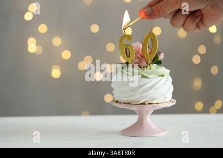 60e anniversaire. Femme allumant des bougies en forme de numéro sur cupcake au bois blanc sur fond gris avec des lumières floues, gros plan. Espace pour le texte Banque D'Images