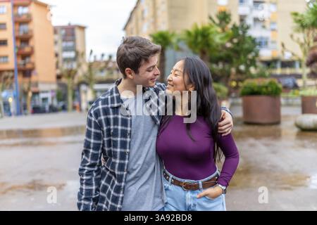 Heureux couple multiethnique partage un moment joyeux à l'extérieur, embrassant et souriant les uns aux autres, entouré d'un cadre urbain avec de la verdure Banque D'Images