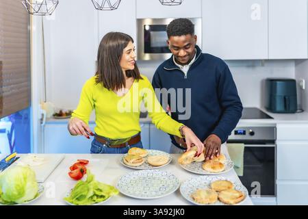 Jeune couple multiethnique préparant des hamburgers pour le dîner, profitant de leur temps ensemble dans une cuisine moderne Banque D'Images
