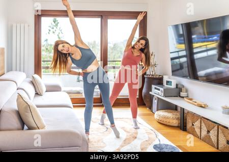 Deux femmes souriantes pratiquant le yoga ensemble dans leur salon, suite à un cours en ligne sur leur télé Banque D'Images