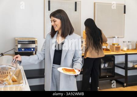 Femme d'affaires souriante tout en choisissant la nourriture du buffet petit déjeuner à l'hôtel pendant le voyage d'affaires, en profitant d'une tranche de pain grillé avec de la confiture Banque D'Images