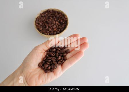 Grains de café torréfiés dans une main avec un bol en osier doux et concentré derrière. Lumière naturelle, fond neutre. Banque D'Images