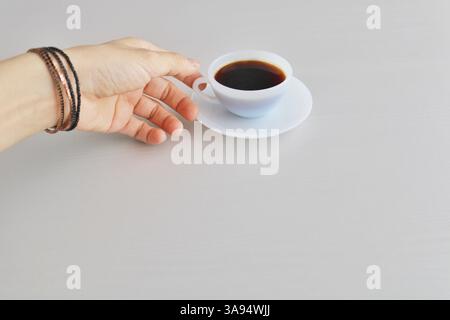 Main tendue pour une tasse de café noir sur une surface propre et neutre. Moment matinal minimal et paisible. Banque D'Images