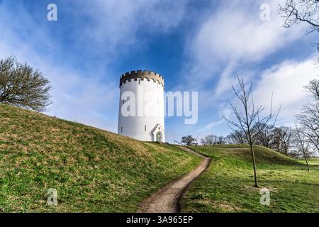 Une scène sereine avec une petite tour blanche entourée de grands arbres sans feuilles, debout sur une colline herbeuse sous un ciel bleu vif. Banque D'Images