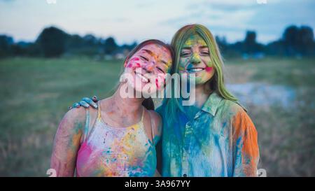 Deux jeunes femmes, couvertes de peinture en poudre holi vibrante, embrassent et sourient, célébrant l'amitié et la joie du festival Banque D'Images