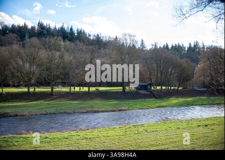 Un paysage tranquille avec une rivière qui coule entouré de berges herbeuses. De l'autre côté, une cabane bleue est nichée parmi les arbres. La colline boisée en t Banque D'Images