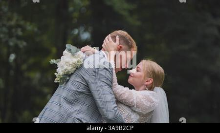 Couple de jeunes mariés partageant un moment tendre, embrassant dans une zone boisée, avec la mariée tenant un bouquet Banque D'Images