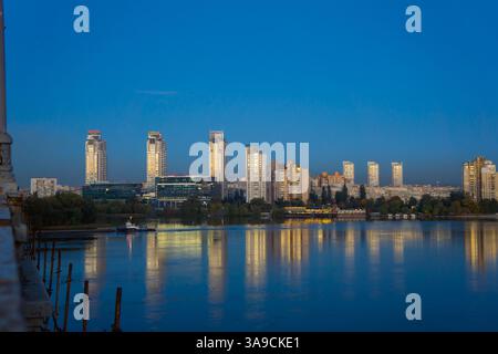 Un paysage urbain moderne au crépuscule avec des bâtiments illuminés reflétant sur l'eau calme des arbres silhouettés au premier plan contrastant avec des structures éclairées Banque D'Images