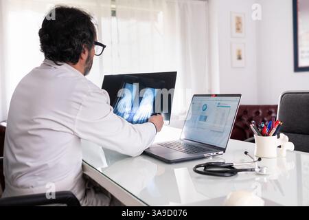 Un médecin d'âge moyen portant une blouse blanche examine une radiographie. Sur son ordinateur portable, il y a un logiciel générique anonyme de soins de santé. En arrière-plan, un autre Banque D'Images